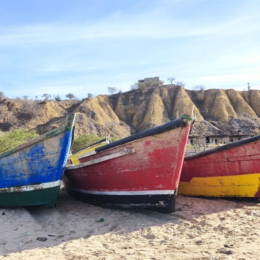 Drei bunte Fischerboote am Strand vor erodierten Sandhügeln unter blauem Himmel.