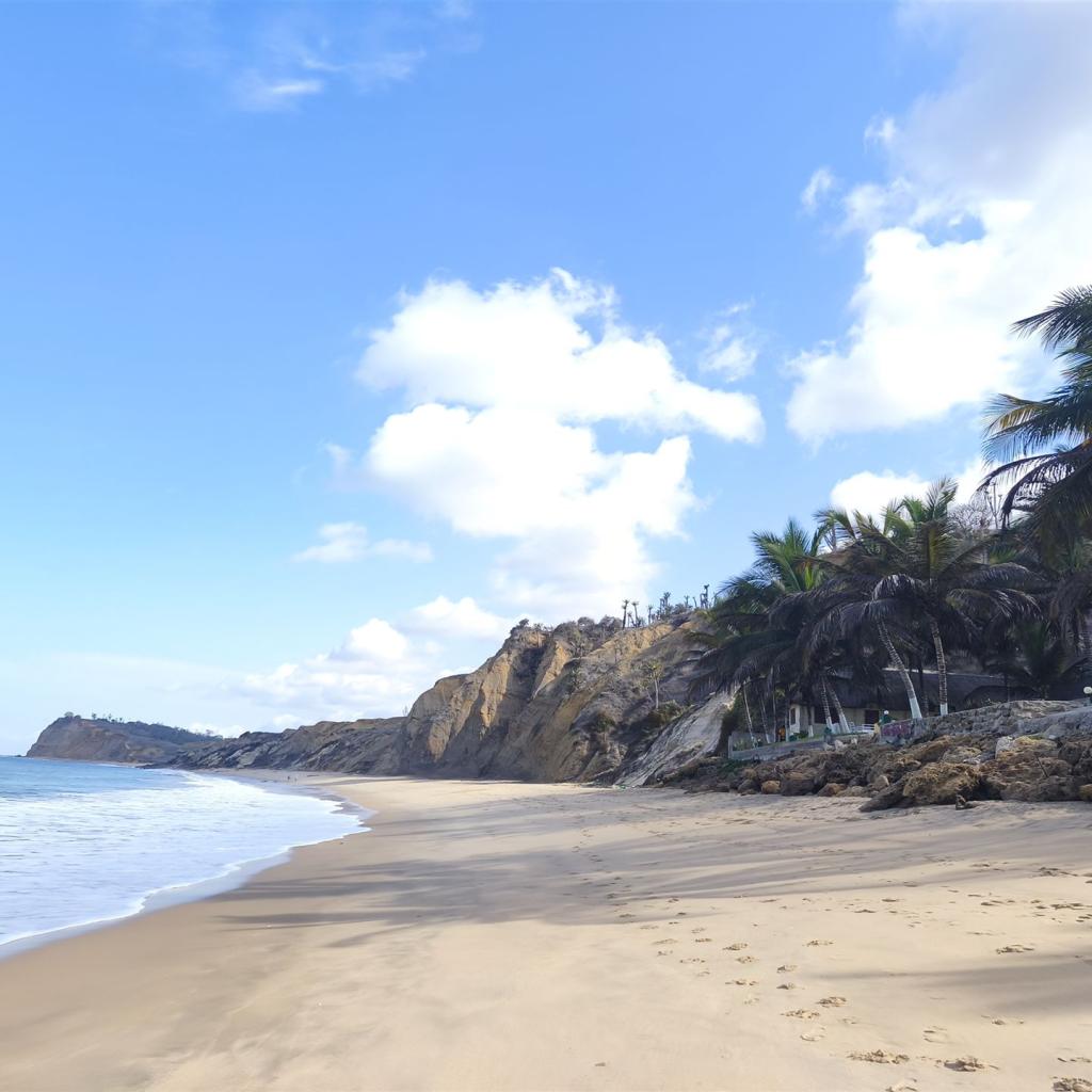 Sonniger Sandstrand mit Palmen, Felsen, Meer und blauem Himmel mit Wolken.
