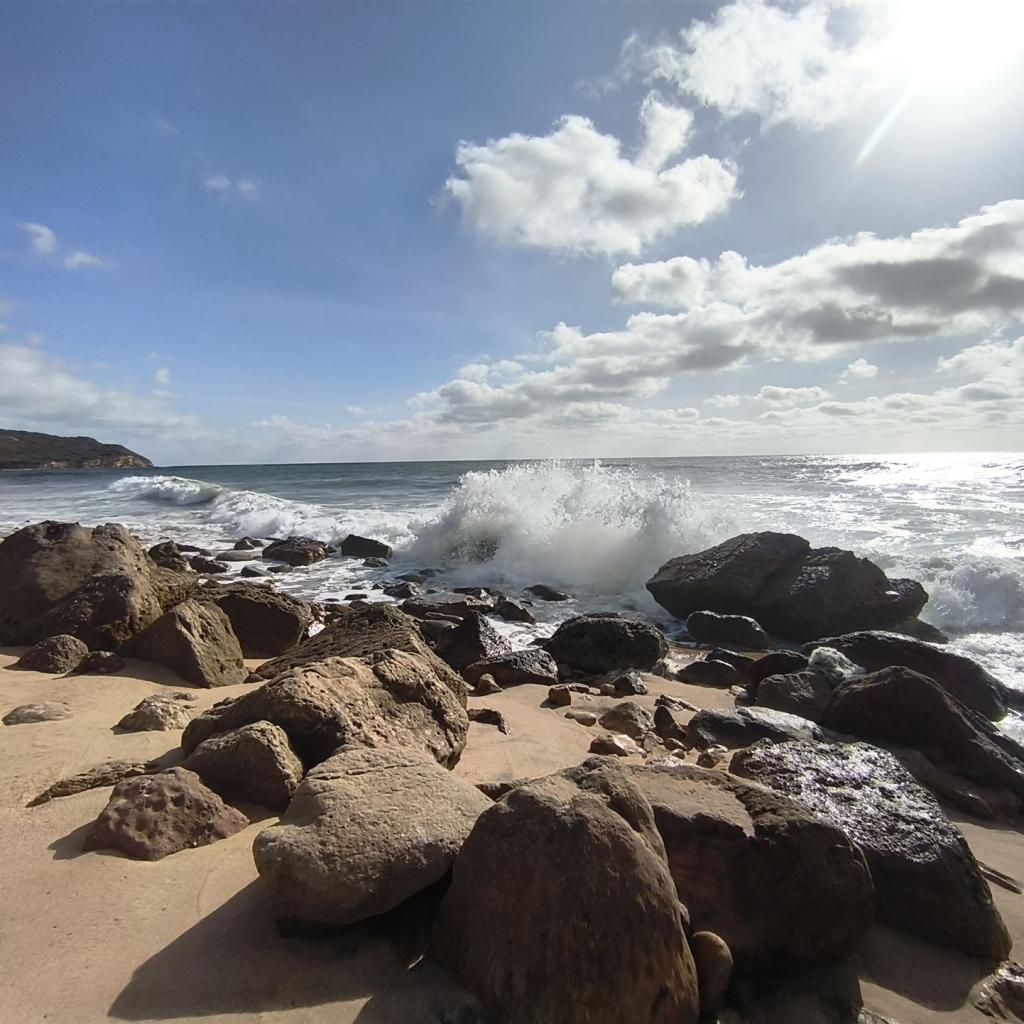 Sonnenbeschienener Strand mit Felsen und krachenden Wellen unter bewölktem Himmel.