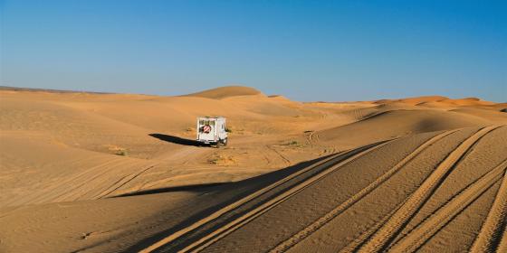 Weite Sanddünengebiete mit einem kleinen weißen Fahrzeug und Fahrspuren im Vordergrund.