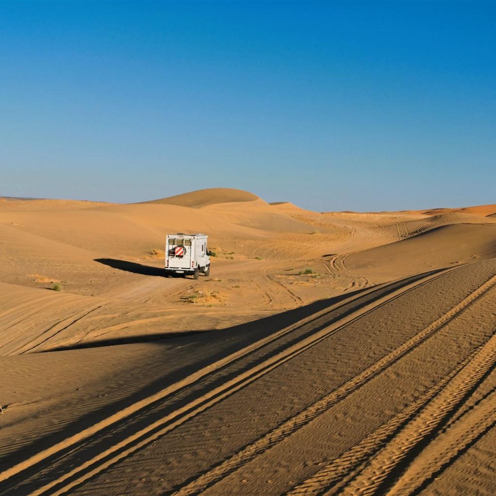 Weite Sanddünengebiete mit einem kleinen weißen Fahrzeug und Fahrspuren im Vordergrund.